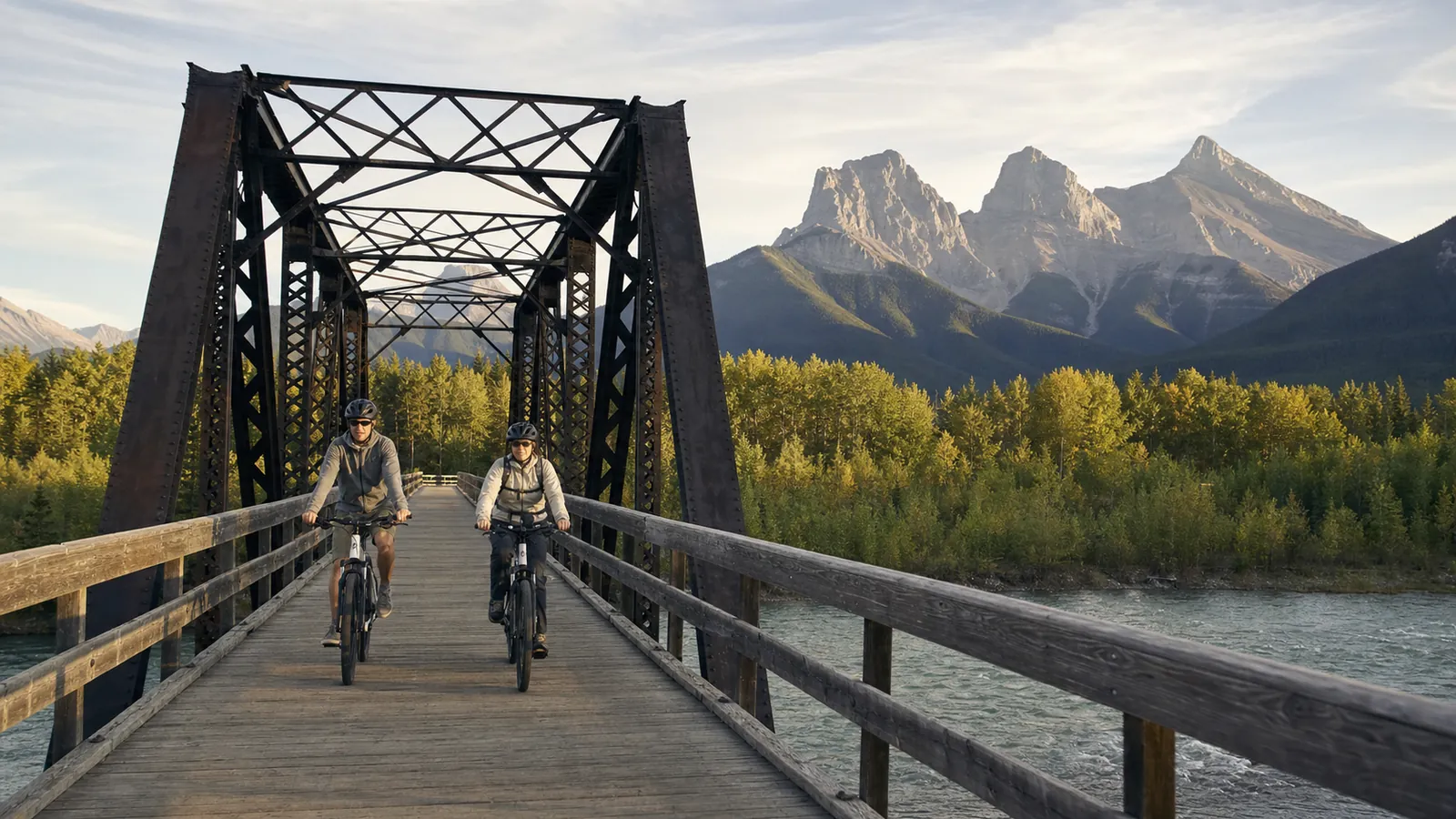 Two e-bike riders crossing Canmore's historic Engine Bridge with the Three Sisters beyond