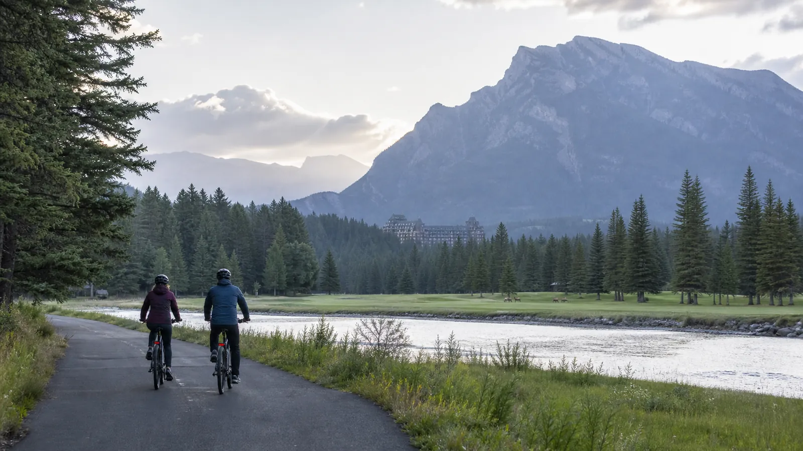 Two e-bike riders beside the Bow River on Golf Course Road with Banff Springs in the distance